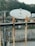 A large satellite dish is positioned in an outdoor area surrounded by trees and a stone wall. The dish is mounted on a structure with metal beams and wires. The foreground includes a rusted metal railing, and there's equipment visible near the wall, suggesting an industrial or telecommunications context.