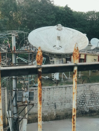 A large satellite dish is positioned in an outdoor area surrounded by trees and a stone wall. The dish is mounted on a structure with metal beams and wires. The foreground includes a rusted metal railing, and there's equipment visible near the wall, suggesting an industrial or telecommunications context.