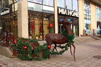 A life-sized decorative reindeer made of wicker stands outside a store named 'MIA HOME'. The reindeer is adorned with green garlands and large red ornaments, resembling a festive Christmas display. The store has glass windows, showing glimpses of more holiday decorations inside, including wreaths and other decorative elements. The surroundings are paved with patterned tiles typical of a pedestrian walkway.