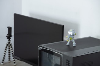 A small, cute robot toy is placed on top of a black computer tower. Behind the computer tower, there is a black monitor. To the left, there is a mini tripod with flexible legs.