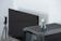 A small, cute robot toy is placed on top of a black computer tower. Behind the computer tower, there is a black monitor. To the left, there is a mini tripod with flexible legs.