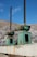 Two large green industrial bolts and brackets are mounted on a concrete structure with a mountainous landscape in the background. The metal appears weathered and slightly rusted. The sky is clear, with a few wispy clouds.