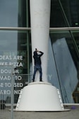 A person dressed in a black jacket and blue jeans stands on a large, white structural base while taking a photograph. The background features a modern glass building with visible text partially reading 'IDEAS ARE LIKE PIZZA.'