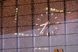 A large, illuminated clock with bright markers and hands is visible against a background of reflective glass windows. The glass windows reflect numerous tiny, warm-colored lights, creating a festive and elegant ambiance. The clock's details are sharply focused, with a modern design.
