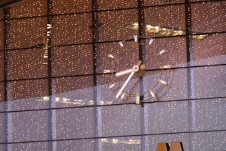 A large, illuminated clock with bright markers and hands is visible against a background of reflective glass windows. The glass windows reflect numerous tiny, warm-colored lights, creating a festive and elegant ambiance. The clock's details are sharply focused, with a modern design.