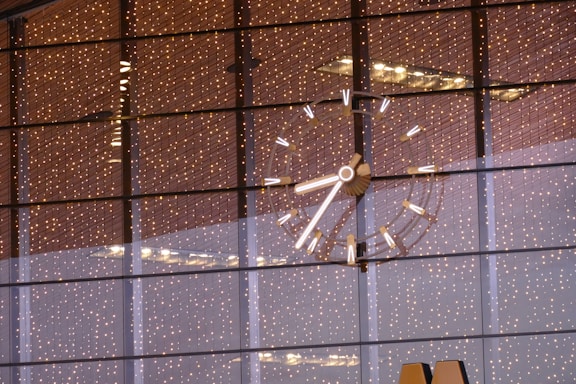 A large, illuminated clock with bright markers and hands is visible against a background of reflective glass windows. The glass windows reflect numerous tiny, warm-colored lights, creating a festive and elegant ambiance. The clock's details are sharply focused, with a modern design.