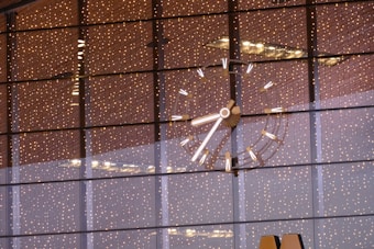 A large, illuminated clock with bright markers and hands is visible against a background of reflective glass windows. The glass windows reflect numerous tiny, warm-colored lights, creating a festive and elegant ambiance. The clock's details are sharply focused, with a modern design.