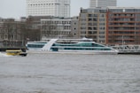 A river scene featuring a sleek, modern white boat with blue-tinted windows cruising past, accompanied by a small yellow water taxi. The background consists of urban architecture with a mix of beige and red brick buildings and some high-rise structures. Bare trees line the embankment, hinting at a cold season.