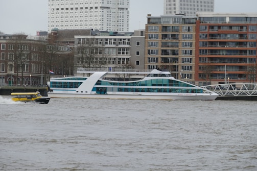A river scene featuring a sleek, modern white boat with blue-tinted windows cruising past, accompanied by a small yellow water taxi. The background consists of urban architecture with a mix of beige and red brick buildings and some high-rise structures. Bare trees line the embankment, hinting at a cold season.