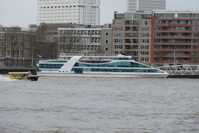 A river scene featuring a sleek, modern white boat with blue-tinted windows cruising past, accompanied by a small yellow water taxi. The background consists of urban architecture with a mix of beige and red brick buildings and some high-rise structures. Bare trees line the embankment, hinting at a cold season.