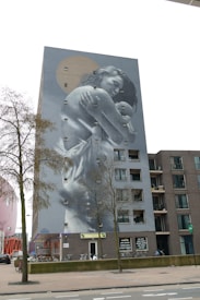 A large mural on the side of an apartment building depicts a mother holding a child. The artwork is monochromatic with a touch of sepia, integrating the building's windows into the design. A tree stands in front, partially obscuring the lower section of the mural. The building is surrounded by a few bicycles and minimal street activity.