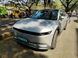 A modern car is parked on a tree-lined street. The vehicle is a Hyundai model, featuring sleek lines and a silver-gray color. In the background, there are shops with colorful signs, several parked motorcycles, and a few people walking, including one individual on the right side of the frame. The street is shaded by large trees with spreading branches, creating a dappled light effect on the ground.