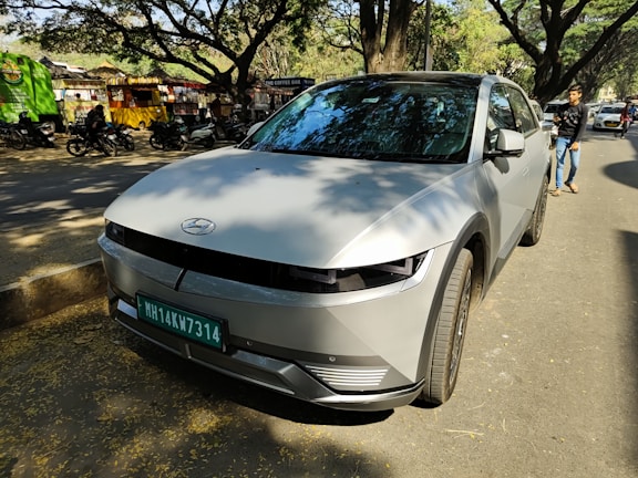 A modern car is parked on a tree-lined street. The vehicle is a Hyundai model, featuring sleek lines and a silver-gray color. In the background, there are shops with colorful signs, several parked motorcycles, and a few people walking, including one individual on the right side of the frame. The street is shaded by large trees with spreading branches, creating a dappled light effect on the ground.