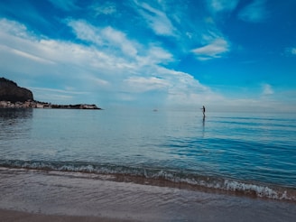 A serene beach scene captures a person paddleboarding on calm ocean waters. In the background, a small sailboat is visible under a sky filled with scattered clouds and shades of blue. A distant shoreline with low-rise buildings and a hill adds depth to the tranquil setting.