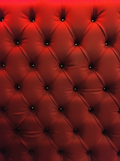 A close-up of a tufted, padded surface with a luxurious texture. The material is upholstered in a deep red color, and small buttons create a diamond pattern.