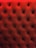 A close-up of a tufted, padded surface with a luxurious texture. The material is upholstered in a deep red color, and small buttons create a diamond pattern.