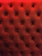 A close-up of a tufted, padded surface with a luxurious texture. The material is upholstered in a deep red color, and small buttons create a diamond pattern.