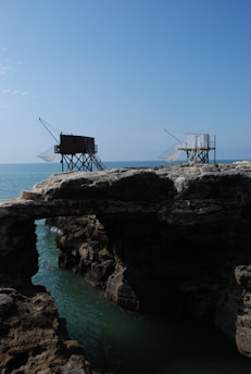 Two elevated fishing huts on stilts with fishing nets are positioned near the edge of rocky cliffs overlooking an expanse of blue ocean. The foreground consists of rugged rocks and a small bridge-like formation with water below.
