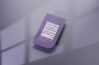 A purple business card with a vertical white text design. The card features one rounded corner and is placed on a reflective, smooth surface. Soft shadows create a subtle depth effect, while a blurred window pattern is faintly visible in the background.