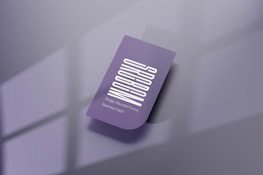 A purple business card with a vertical white text design. The card features one rounded corner and is placed on a reflective, smooth surface. Soft shadows create a subtle depth effect, while a blurred window pattern is faintly visible in the background.