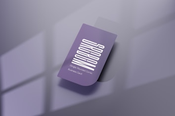 A purple business card with a vertical white text design. The card features one rounded corner and is placed on a reflective, smooth surface. Soft shadows create a subtle depth effect, while a blurred window pattern is faintly visible in the background.