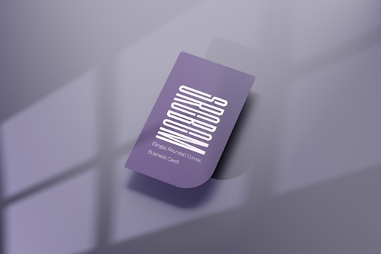 A purple business card with a vertical white text design. The card features one rounded corner and is placed on a reflective, smooth surface. Soft shadows create a subtle depth effect, while a blurred window pattern is faintly visible in the background.