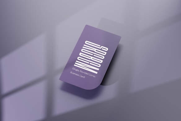 A purple business card with a vertical white text design. The card features one rounded corner and is placed on a reflective, smooth surface. Soft shadows create a subtle depth effect, while a blurred window pattern is faintly visible in the background.