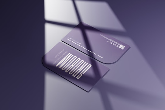 Two sleek, rounded-edge business cards are placed on a smooth surface, casting shadow patterns reminiscent of window panes. The cards are predominantly purple with modern, minimalist typography. One card is facing up, displaying contact information and a QR code, while the other card has graphic elements.