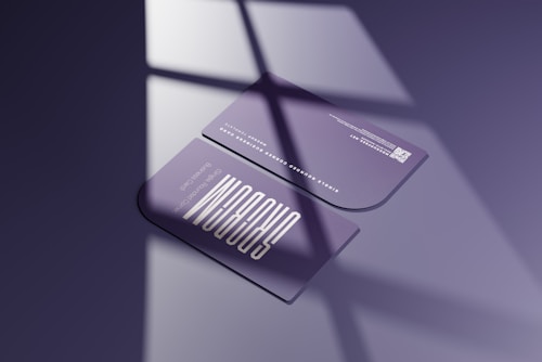 Two sleek, rounded-edge business cards are placed on a smooth surface, casting shadow patterns reminiscent of window panes. The cards are predominantly purple with modern, minimalist typography. One card is facing up, displaying contact information and a QR code, while the other card has graphic elements.