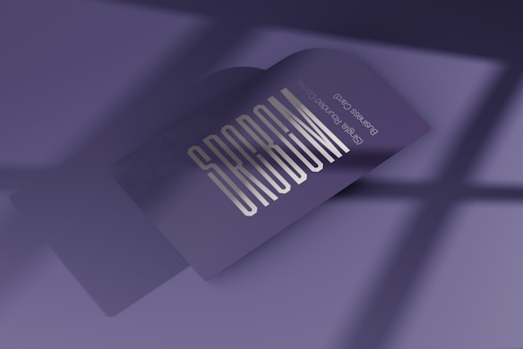 A business card with bold, stylized text reading 'SPREAD' is prominently displayed. The card is in a folded position, casting a soft shadow over a smooth, purple surface. The lighting creates a modern and professional ambiance, with a subtle gradient in the background.