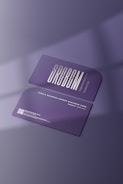 Two purple business cards featuring sleek typography and a single rounded corner are placed on a surface with soft shadows. The foreground card prominently displays the brand name in bold letters, while the background card includes contact details and a QR code in white text.
