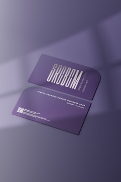 Two purple business cards featuring sleek typography and a single rounded corner are placed on a surface with soft shadows. The foreground card prominently displays the brand name in bold letters, while the background card includes contact details and a QR code in white text.