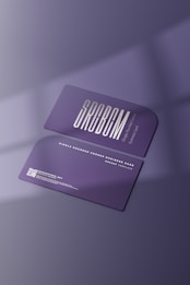Two purple business cards featuring sleek typography and a single rounded corner are placed on a surface with soft shadows. The foreground card prominently displays the brand name in bold letters, while the background card includes contact details and a QR code in white text.