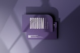 A business card mockup with a rounded corner design placed on a purple surface. The card features bold, modern typography with the text 'SRCBCM' prominently displayed. Light and shadow effects create a stylish and professional atmosphere.