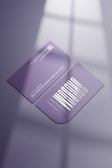 Two purple business cards are placed on a reflective surface. One card is facing up showing a stylized white text and the other is facing down displaying a QR code and details in white. The lighting creates soft shadows and a subtle window reflection on the surface.