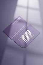 Two purple business cards are placed on a reflective surface. One card is facing up showing a stylized white text and the other is facing down displaying a QR code and details in white. The lighting creates soft shadows and a subtle window reflection on the surface.