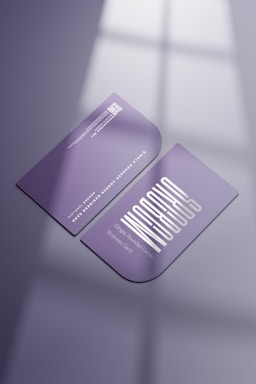 Two purple business cards are placed on a reflective surface. One card is facing up showing a stylized white text and the other is facing down displaying a QR code and details in white. The lighting creates soft shadows and a subtle window reflection on the surface.