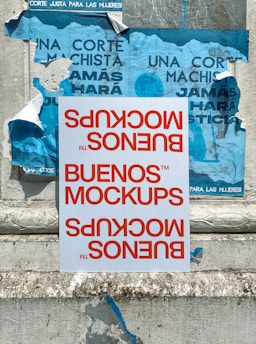 A white paper with red text reading 'BUENOS MOCKUPS' is placed upside down against a weathered wall. The wall is adorned with torn, blue posters featuring Spanish text. The peeling posters suggest age and neglect.