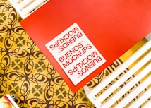 A red tabletop with a document labeled 'BUENOS MOCKUPS' in bold red letters. The table is next to a patterned floor featuring intricate designs in brown and yellow. White slats are seen next to the red table.