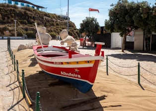 A red and blue wooden boat named Albufeira is placed on sandy ground, surrounded by a low chain link fence. The boat features yellow and blue decorative designs and has two figures made of sand seated inside, resembling fishermen. In the background, there is a hill with greenery, a modern structure with a blue glass facade, and a few trees providing shade.