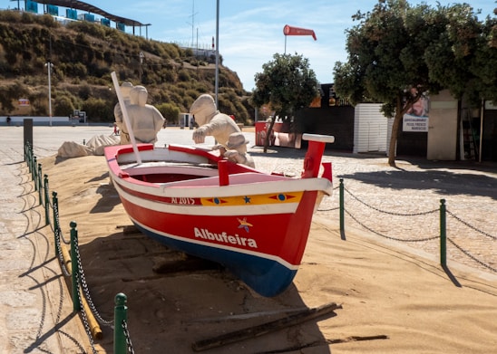 A red and blue wooden boat named Albufeira is placed on sandy ground, surrounded by a low chain link fence. The boat features yellow and blue decorative designs and has two figures made of sand seated inside, resembling fishermen. In the background, there is a hill with greenery, a modern structure with a blue glass facade, and a few trees providing shade.