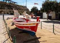A red and blue wooden boat named Albufeira is placed on sandy ground, surrounded by a low chain link fence. The boat features yellow and blue decorative designs and has two figures made of sand seated inside, resembling fishermen. In the background, there is a hill with greenery, a modern structure with a blue glass facade, and a few trees providing shade.