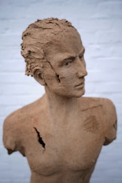 A detailed clay sculpture of a human bust, featuring realistic facial features and a textured surface. The sculpture has noticeable cracks and divots, adding to its aged appearance. It is positioned against a plain, light-colored background.