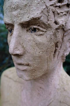 A close-up view of a clay sculpture depicting a human face, showing intricate details in the texture such as cracks and rough surfaces. The eyes are dark and slightly inset, giving the sculpture a contemplative expression.