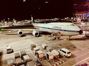 An airplane from Lufthansa is parked on the tarmac of an airport at night. The scene includes various airport ground vehicles and equipment scattered around. The background features a well-lit terminal and other airport facilities.