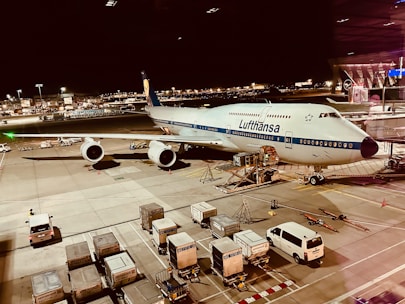 An airplane from Lufthansa is parked on the tarmac of an airport at night. The scene includes various airport ground vehicles and equipment scattered around. The background features a well-lit terminal and other airport facilities.