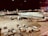An airplane from Lufthansa is parked on the tarmac of an airport at night. The scene includes various airport ground vehicles and equipment scattered around. The background features a well-lit terminal and other airport facilities.