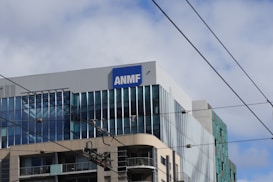 A modern multi-story building with a sign labeled ANMF near the top. The building features large glass windows with reflective surfaces. There are visible overhead power lines crossing the image.