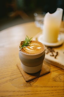 A cup of coffee with latte art sits on a wooden table. The cup is placed on a small wooden coaster and has a decorative sprig of rosemary resting on its rim. In the background, there is a large candle on a ceramic plate and scattered coffee beans.
