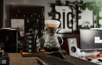A cozy workspace featuring a pour-over coffee setup with a glass carafe and black kettle on a wooden tray. The background includes a blurred monitor and speakers, along with green foliage and framed artwork, creating a warm and inviting atmosphere.